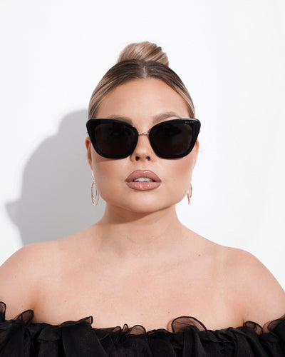 sunglasses, kyst eye, cats eye silhouette, black frame, wide face sunglasses, shown as worn by a woman wearing her blond hair tied back in a slick bun, gold earrings, and a black ruffled off the shoulder top
