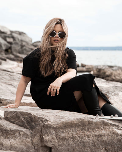 sunglasses, kyst eye, cat eye silhouette, white frame, smokey lens, wide face sunglasses, shown as a worn by a women with long blond hair, a black dress and black boots, she is sitting on rocks near the water